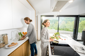 Young cheerful couple dressed alike in gray sweaters cooking some veggie food at the modern kitchen