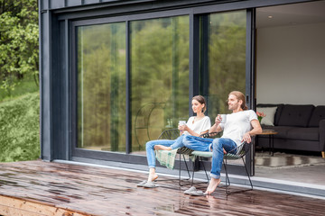 Young couple sitting with cups on the terrace of the modern house enjoying beautiful view outdoors