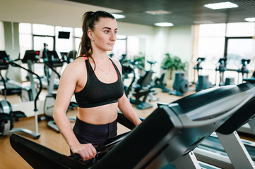 Fototapeta premium Beautiful girl is engaged fitness in sport club. Attractive young woman running on a treadmill. Fitness trainer is engaged in sports in gym. Side view.