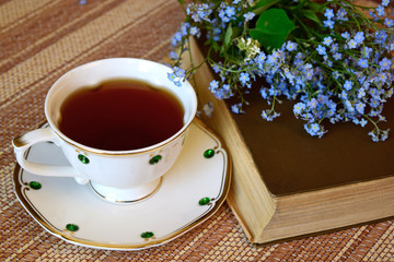 On the table a book, a bouquet of flowers and a Cup of tea. The ancient book is a bouquet of forget-me-nots. An old brown book and a white Cup of tea.