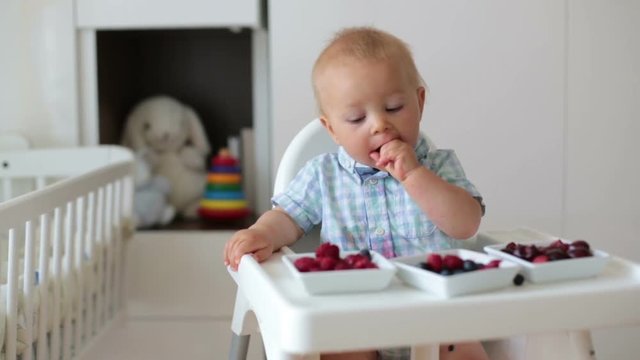 Adorable little baby boy, eating fresh fruits at home, sitting in baby chair in kids room