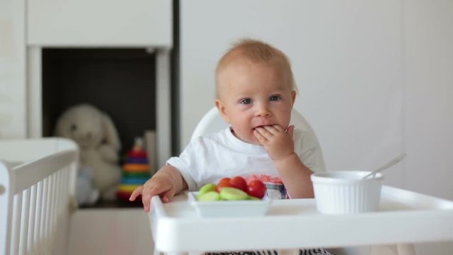 Sweet baby child, boy, eating mashed food and fresh vegetables, sitting in high baby chair in kids room