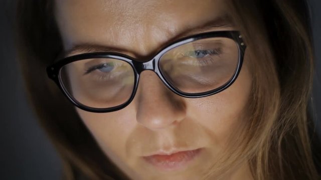 Woman is browsing news or social information feed online with her laptop, smartphone or tablet device. Monitor screen reflections could be seen in her eyeglasses glass.