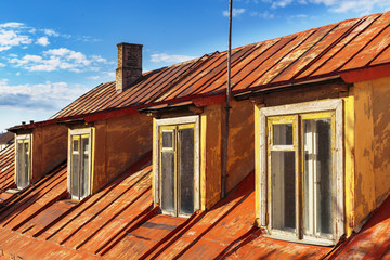 Old rusted metal red mansard roof with four wooden windows and brick chimney on blue sky background