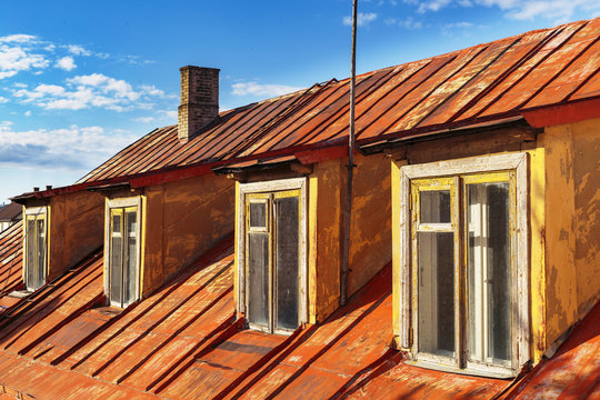 Old Rusted Metal Red Mansard Roof With Four Wooden Windows And Brick Chimney On Blue Sky Background