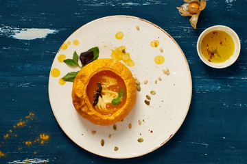 Delicious vegetable soup decorated with basil leaves and cooked in pumpkin lying on flat round plate standing on blue aged wooden surface. Tasty restaurant vegetarian meal, veggie recipe. Top view.