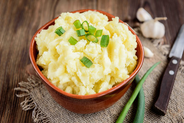Mashed potatoes in bowl on wooden table with spring green onion and garlic.