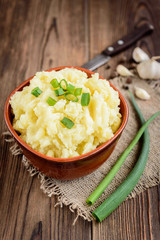 Mashed potatoes in bowl on wooden table with spring green onion and garlic.