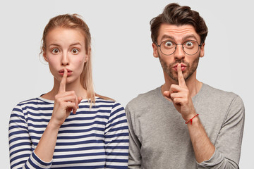 Amazed European female and male make silence sign, keep fore fingers on lips, tell secret and...