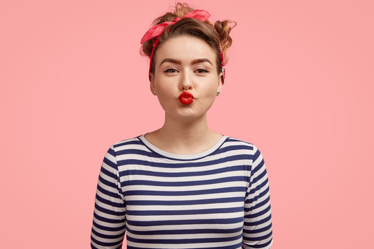 Beautiful young female gives kiss at camera, shows brightly red lips, has make up, wears retro style clothing, poses against pink background. Charming pinup girl in striped sweater flirts with someone