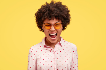 Fototapeta na wymiar Annoyed African American female freaks out and excliams angrily, being depressed as has many work and deadlines, wears stylish clothes, isolated over yellow background. Negative emotions concept