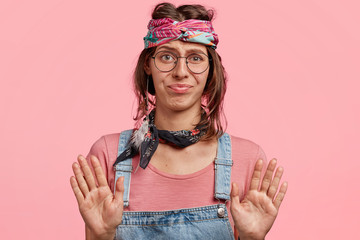 Beautiful hippie girl poses against pink background, makes stop gesture, has displeased expression,...