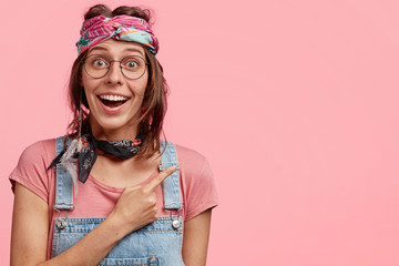 Happy adorable hippie woman indicates aside with cheerful expression, shows positive attitude, poses against pink background, shows nice cafe for spending leisure time. Friendly hippy girl indoor