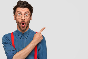 Surprised male hipster, keeps mouth opened, being stupefied as advertises something, wears fashionable shirt and red suspenders, indicates with fore finger at upper right corner. Omg concept - obrazy, fototapety, plakaty