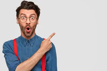 Deurstickers Surprised male hipster, keeps mouth opened, being stupefied as advertises something, wears fashionable shirt and red suspenders, indicates with fore finger at upper right corner. Omg concept © Wayhome Studio
