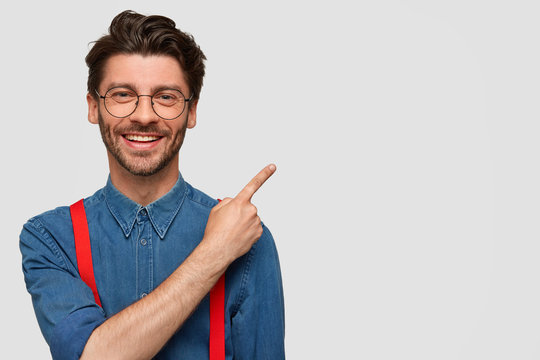 Cheerful pleasant looking young male with bristle, has good mood, smiles pleasantly, points with fore finger at blank space, advertises new products at shop, stands against white background.
