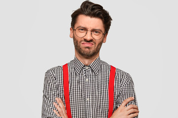 Dissatisfied unshaven male designer keeps hands crossed, frowns face, being discontent with new designing project, wears checkered shirt, sees something unpleasant, isolated over white wall.