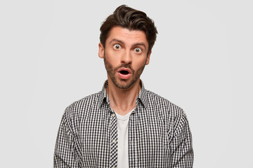 Indoor shot of stunned European unshaven male keeps mouth opened, wears stylish checkered shirt, feels surprisement while watches excited detective programme, isolated over white studio wall.