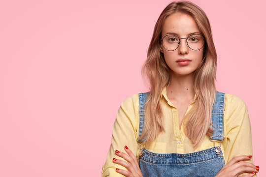 Studio sho of pleased attractive female student wears glasses, yellow shirt and denim overalls, keeps arms folded, has break after classes, poses against pink background with blank space on left