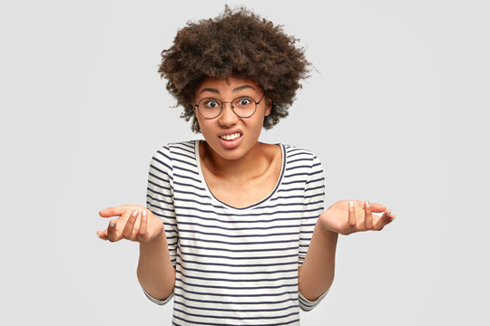 Hesitant African American female models in studio, has puzzled expression, shruggs shoulders with uncertainty, frowns face. Doubtful woman costumer can`t make choice, isolated on white wall.
