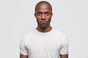Photo of serious dark skinned male with surprised expression, feels astonishment as sees something unexpected, wears casual t shirt, isolated over white background. People and facial expressions