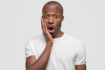 Emotional African American guy keeps hand on cheek, mouth opened, looks in bewilderment at camera, feels shock after failure, stands against white background. People, ethnicity, emotions concept