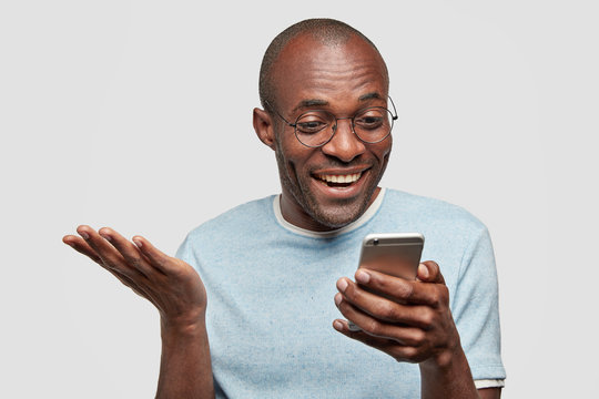 Happy middle aged African Amerfican male holds modern smart phone, recieves good message on cellular, reads news in internet, has plesant smile, keeps hand raised, poses indoor. Technology concept