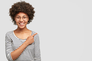 Horizontal portrait of happy dark skinned female with Afro hairsyle giggles joyfully, has broad smile with white teeth, indicates aside at blank space for your advertising content. Emotions concept