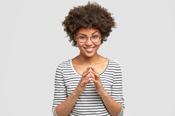 Attractive African American female with curly dark hair, keeps hands together and looks with intrigue, wears casual striped jacket and glasses poses in studio. Cheerful housewife anticipates something