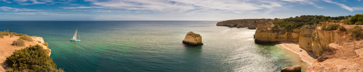 Panorama of the Algarve coastline in Portugal with a sailing boat moving towards the Marinha beach. There are cliffs, vegetation, a beach and rock stacks in the scene.