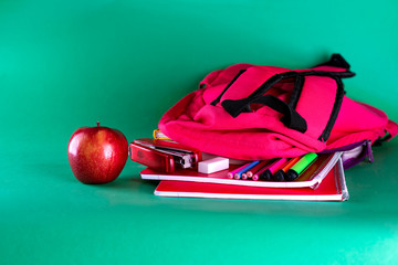 Back to school with health food. Backpack with school supplies and red apple on green background. Healthy lifestyle concept.