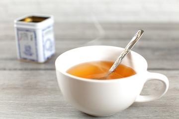 Cup of hot amber tea with steam, with metal tin placed on gray wood desk in background.