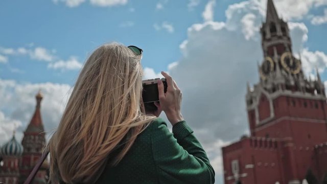 Blonde young tourist in a green coat making photos of The Spasskaya Tower, the main tower on the eastern wall of the Moscow Kremlin. The Red Square. Summer day. Slider real time medium shot.