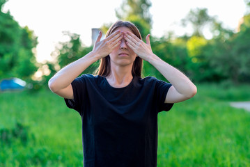 The brunette girl in summer in a park nature. Gestures of the hands closed her eyes. Peekaboo. Emotional gestures I do not see. In everyday clothes.