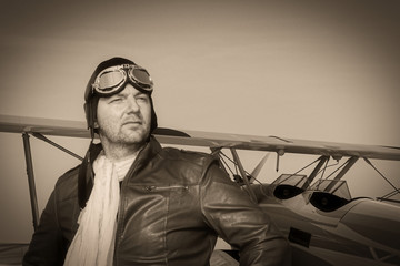 Portrait of a vintage pilot with leather cap, scarf and aviator glasses in front of a historic airplane biplane - Portrait of a man in historical pilot clothing - vintage old picture style
