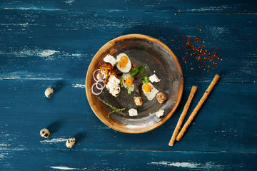 Bruschetta with cream cheese, fried eggs, mushrooms and salad leaves lying on plate standing on blue aged wooden surface with quail eggs and bread sticks scattered around. Tasty daily meal. Top view.