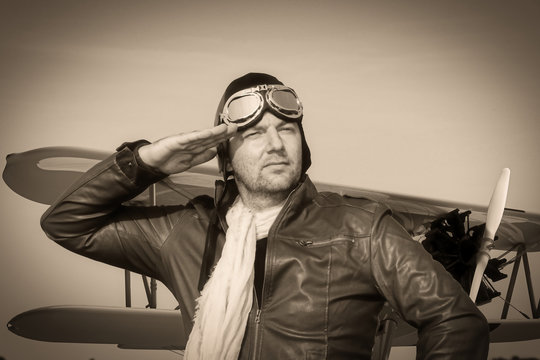 
Portrait of a vintage pilot with leather cap, scarf and aviator glasses salutes in front of a historical biplane - Portrait of a man in historical pilot clothing - vintage old picture style