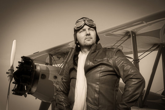 Portrait of a vintage pilot with leather cap, scarf and aviator glasses in front of a historic airplane biplane - Portrait of a man in historical pilot clothing - vintage old picture style
