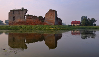 Ruiny średniowiecznego zamku, obok współczesny dom, ruiny na zielonej grobli, w wodzie piękne lustrzane odbicie
