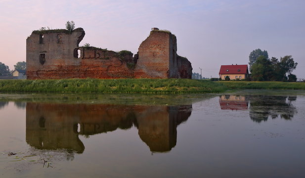 Ruiny średniowiecznego zamku, obok wsp&oacute;łczesny dom, ruiny na zielonej grobli, w wodzie piękne lustrzane odbicie