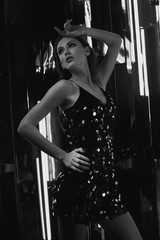 Brunette woman wearing elegant black sequin strap cami dress standing on dark interior with mirrors. Attractive caucasian female model posing against glowing neon lamps and mirrors on background