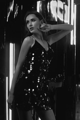 Brunette woman wearing elegant black sequin strap cami dress standing on dark interior with...