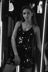 Brunette woman wearing elegant black sequin strap cami dress standing on dark interior with mirrors. Attractive caucasian female model posing against glowing neon lamps and mirrors on background