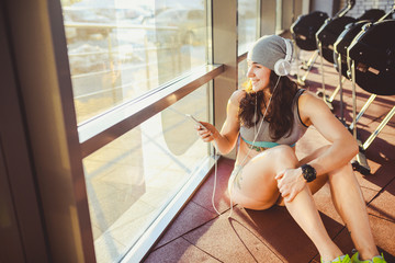 Theme sports, health and technology. beautiful sexy Caucasian woman sportswoman in gray sportswear and hat sits by window with sun setting uses smartphone listen music in large headphones on head