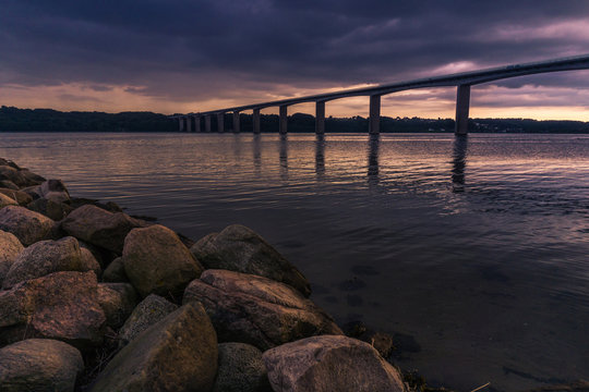 View at the bridge named Vejlefjordbroen and its reflections from Vejle, Denmark. Big stones in the foreground and a beautiful sunset and moody clouds.