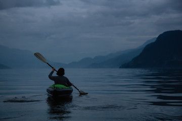 Adventurous woman kayaking during a dark gloomy evening surrounded by Canadian Mountain Landscape. Taken in Howe Sound, North of Vancouver, BC, Canada.