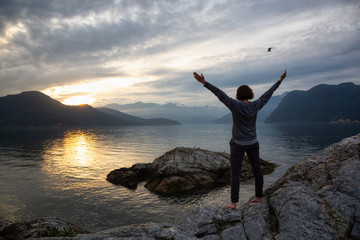 Woman enjoying the beautiful Canadian Mountain Landscape during a vibrant sunset. Taken on a rocky Island in Howe Sound near Vancouver, BC, Canada.