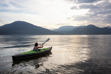 Woman kayaking around the beautiful Canadian Mountain Landscape during a vibrant cloudy evening. Taken in Howe Sound, North of Vancouver, British Columbia, Canada.