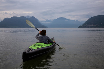 Woman kayaking around the beautiful Canadian Mountain Landscape during a vibrant cloudy evening. Taken in Howe Sound, North of Vancouver, British Columbia, Canada.