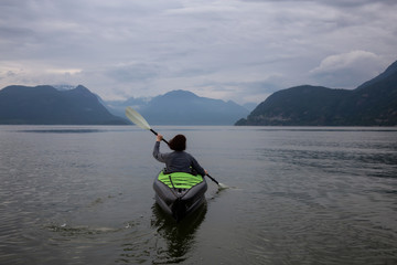 Woman kayaking around the beautiful Canadian Mountain Landscape during a vibrant cloudy evening. Taken in Howe Sound, North of Vancouver, British Columbia, Canada.